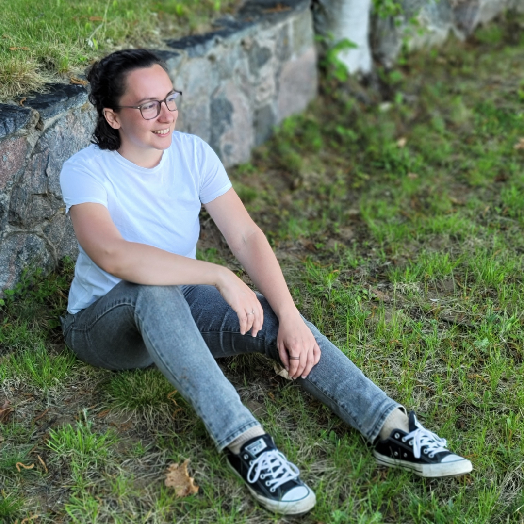 a photo of Eva in a white tshirt, grey jeans and convers, sitting on grass and looking away from the camera, smiling