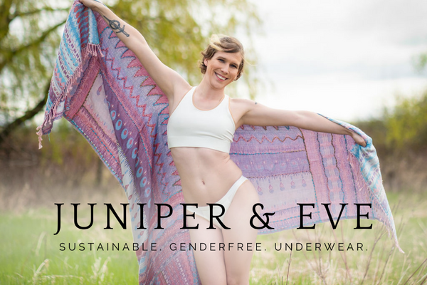A photo of a pretty trans woman in white underwear standing outdoors, holding a colourful shawl behind her, with the Juniper & Eve logo superimposed in black over the lower portion of the image